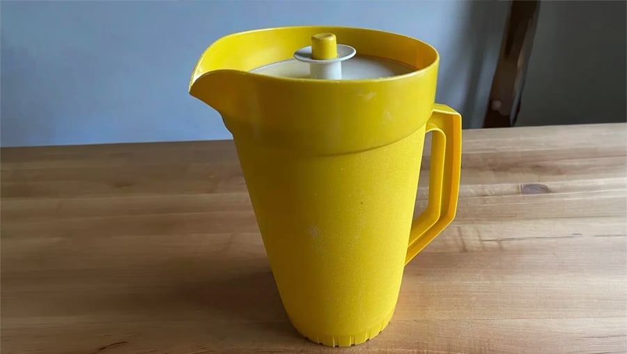 A yellow plastic pitcher with a lid sits on a wooden table. The pitcher has a handle and a spout, and the background is slightly out of focus.