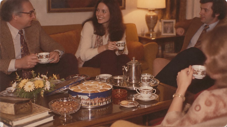 Four adults sit in a cozy living room, smiling and drinking tea. A coffee table holds a tin of cookies, tea set, almonds, and flowers. The mood is warm and friendly, with soft lighting from a table lamp.