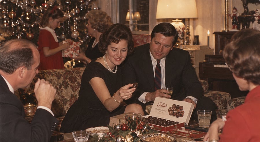 People dressed in formal attire sit around a living room table, sharing a box of chocolates. A decorated Christmas tree and festive decorations fill the background, creating a warm holiday atmosphere.