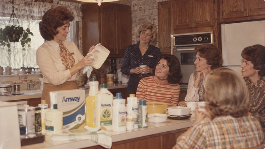 A woman demonstrates an Amway cleaning product to a group of women gathered in a kitchen.