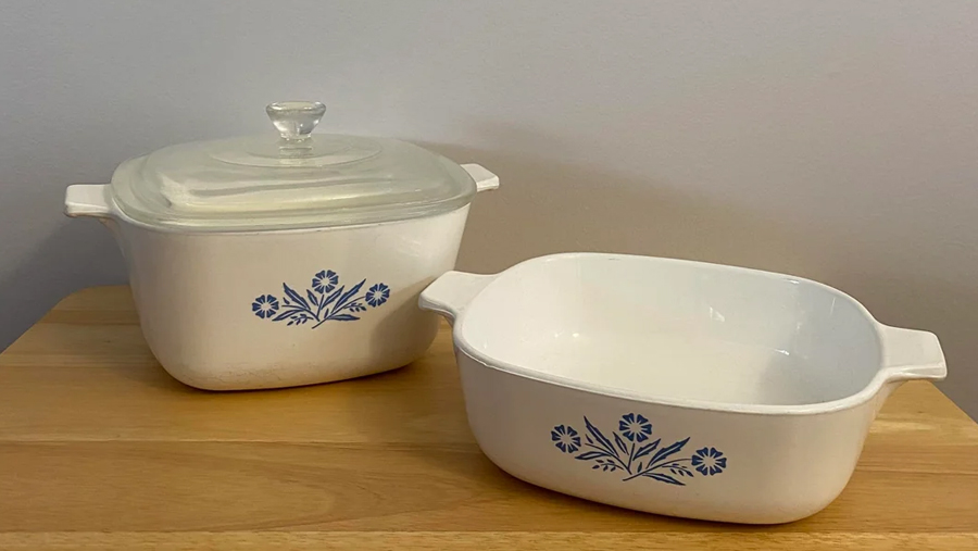 Two white vintage casserole dishes with blue floral designs sit on a wooden surface. One dish has a clear glass lid, while the other is open. Both have handles on the sides.