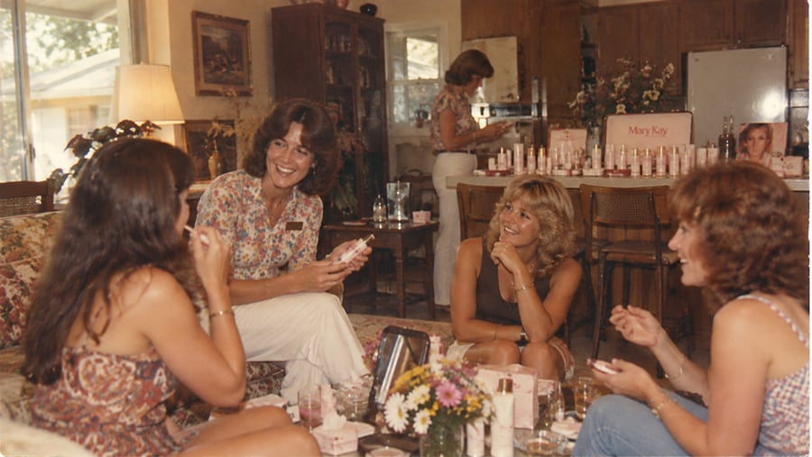 Four women gathered in a living room during a Mary Kay cosmetic demonstration, with various beauty products displayed on a kitchen counter in the background.