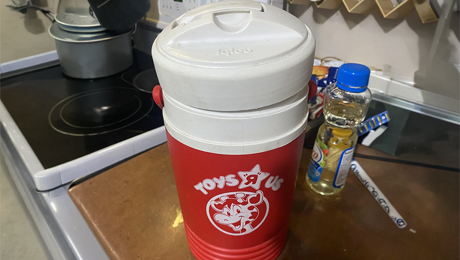 A red and white Toys “R” Us-branded Igloo water cooler sits on a kitchen counter near a bottle of cooking oil and a stovetop.