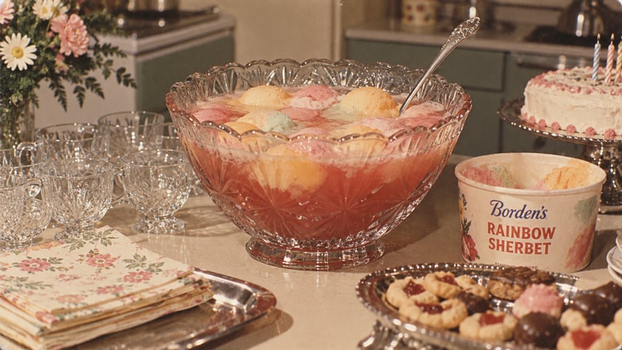 A crystal punch bowl with rainbow sherbet punch sits on a table with matching glasses, floral napkins, cookies, a cake with candles, and a tub of Borden’s Rainbow Sherbet.