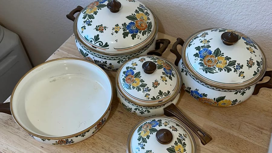 A set of white enamel cookware with floral patterns, including two lidded pots, one lidded saucepan, an empty skillet, and an extra lid, resting on a wooden surface.