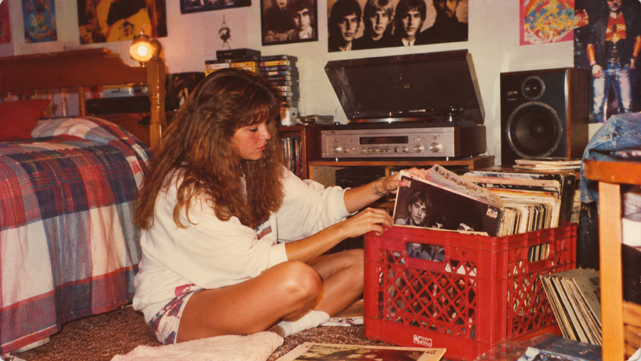 A young woman sits on the floor in a cozy, retro bedroom, flipping through vinyl records in a red crate. Posters, a stereo system, and a checkered bedspread complete the vintage vibe.