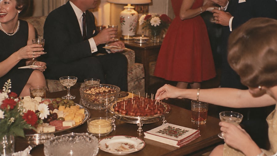 People in formal attire sit around a coffee table with snacks, drinks, and flowers, enjoying a social gathering. One person reaches for an appetizer from a tray.