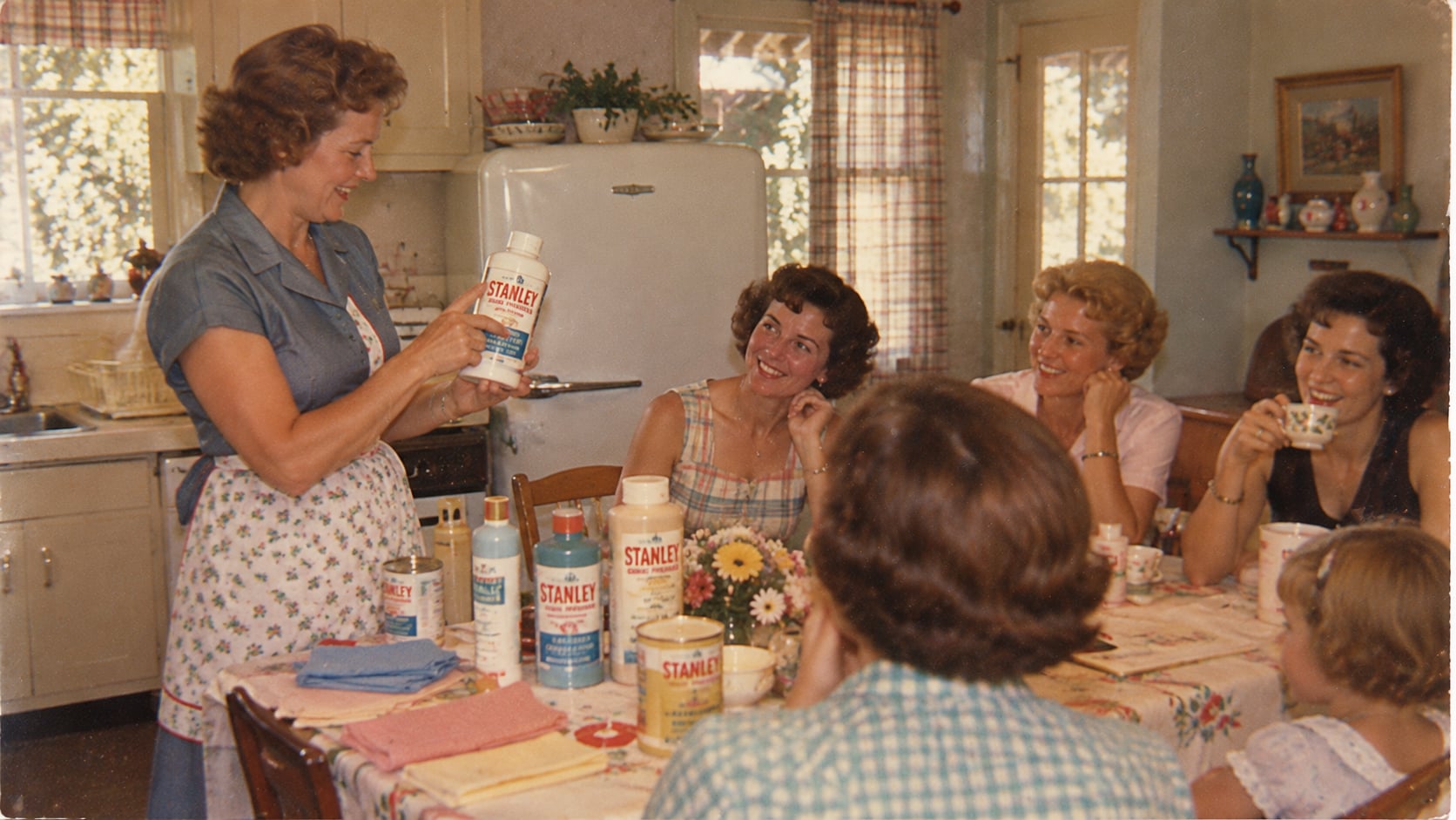 A woman in an apron demonstrates a Stanley cleaning product to four other women and a child sitting around a kitchen table.