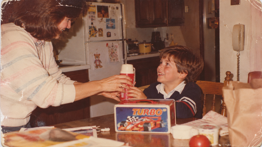 A woman hands a smiling young boy a red thermos at a kitchen table. The boy sits near a Turbo lunchbox. The background shows a fridge with magnets and wood cabinets, suggesting a warm, homey environment.