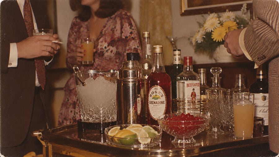Three people stand around a bar cart with various bottles, glasses, and garnishes. Two hold drinks, one holds a cigarette. A vase of yellow flowers is in the background.