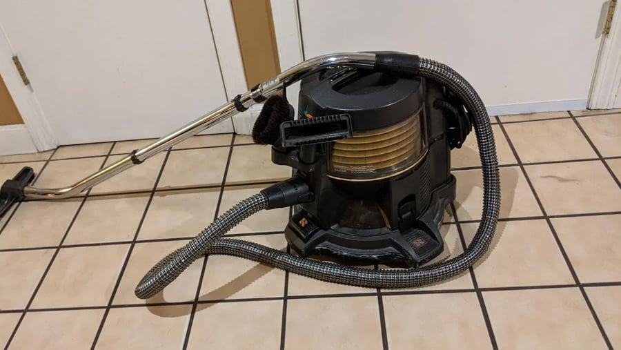Black canister vacuum cleaner with a chrome wand and flexible hose on a tiled floor.