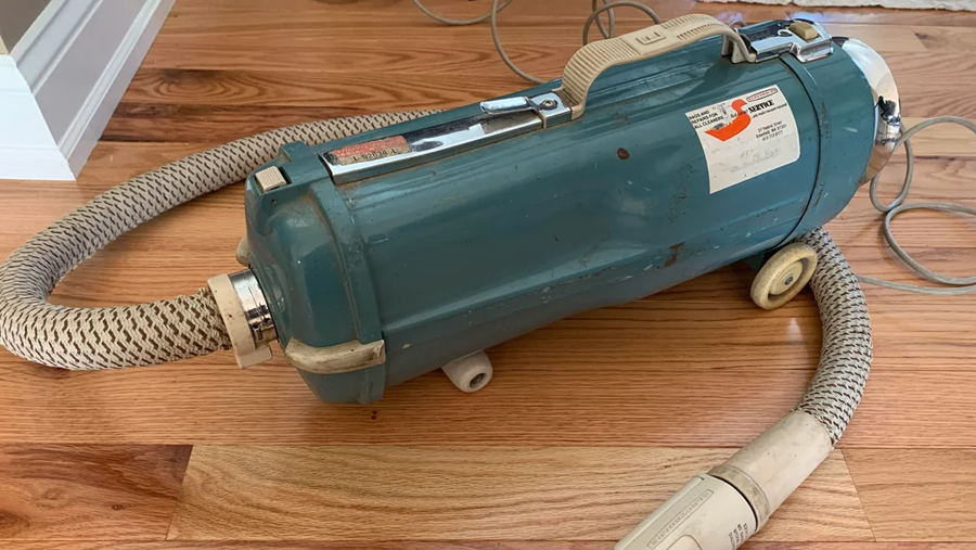 A vintage blue canister vacuum cleaner with a woven hose and beige handle sits on a wooden floor. The vacuum has wheels and an orange and white label on top.