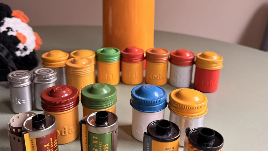 Several colorful Kodak film canisters and rolls are arranged on a table, with a large yellow cylinder and a plush toy in the background. The canisters are red, yellow, orange, green, and silver.