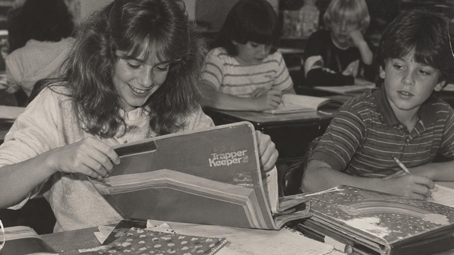 A girl in a classroom smiles while opening a Trapper Keeper folder at her desk. Other students around her are writing and looking at their notebooks.