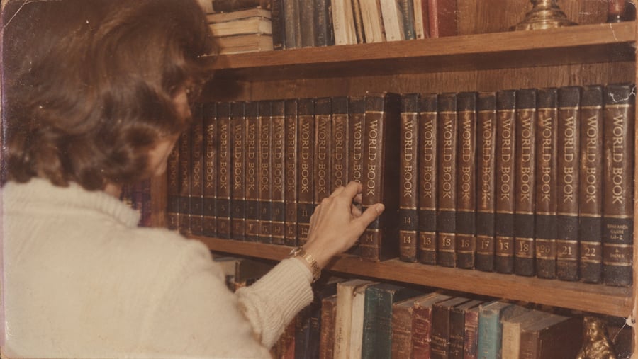 A woman reaches to pull a volume of the World Book encyclopedia from a wooden bookshelf.