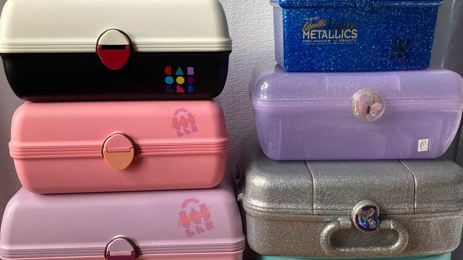 A stack of colorful, vintage Caboodles makeup cases in various colors and finishes, including pink, purple, black, blue, and silver, sit against a white wall.