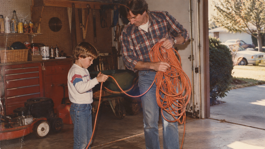 A man and a boy are winding an orange extension cord together in a garage. Tools and a lawnmower are visible, and sunlight streams through the open garage door.