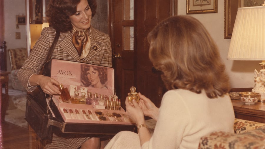 An Avon representative shows a case of cosmetics and perfume to a seated customer.