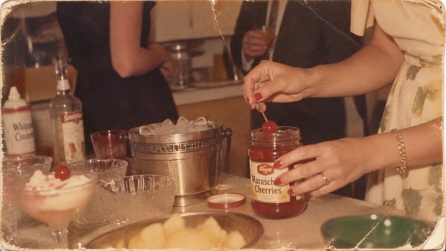 A woman in a floral dress takes a maraschino cherry from a jar at a vintage party, with dessert bowls, whipped cream, and an ice bucket on the counter.