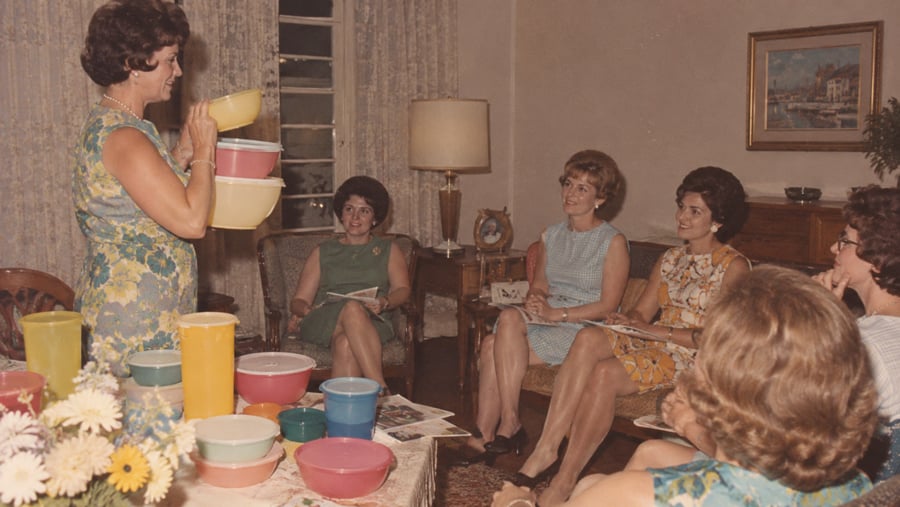 A woman in a floral dress demonstrates plastic bowls to a group of women sitting in a living room.