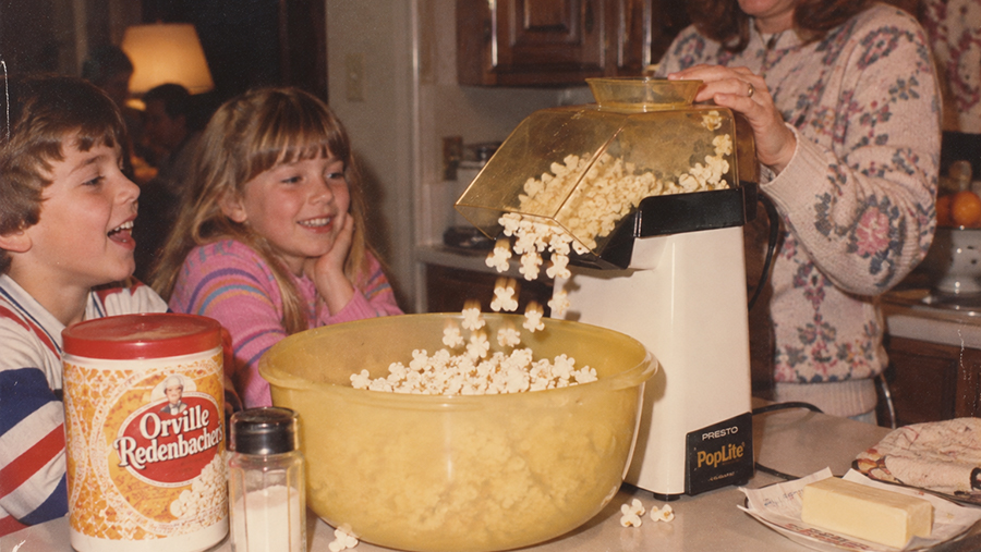 Two smiling kids watch popcorn pop into a large yellow bowl from a popcorn maker. An adult pours the popcorn. A canister of Orville Redenbacher popcorn and salt are on the counter.