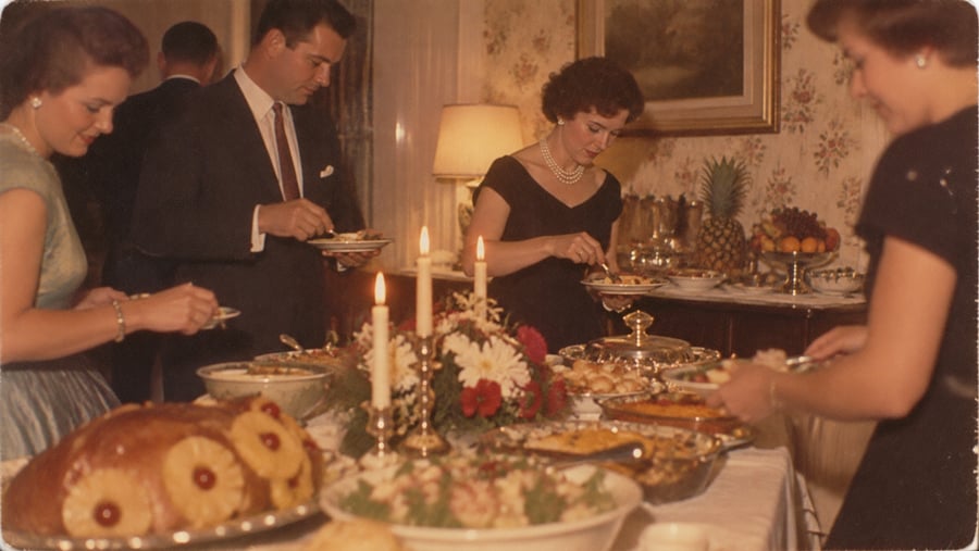 Four well-dressed adults serve themselves food from a buffet table decorated with candles, flowers, and various dishes at a formal gathering in a warmly lit room.
