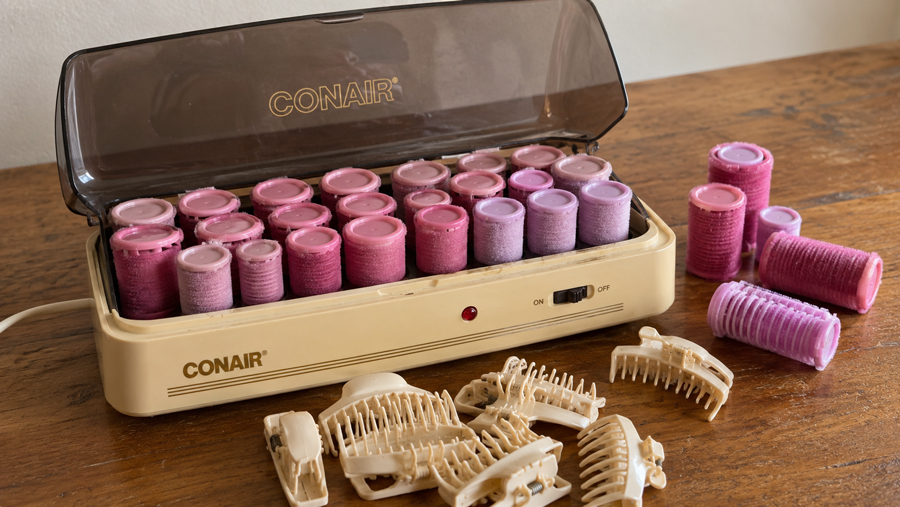 A beige Conair hair roller set with pink and purple rollers heating in the case. Several hair clips and extra rollers are placed on the wooden table next to the set.