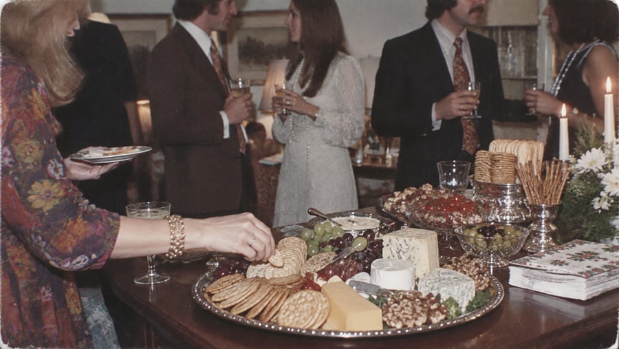People in formal attire stand socializing at a party. A table in the foreground is set with cheese, crackers, fruit, and candles. One person is reaching for food. The atmosphere appears warm and festive.