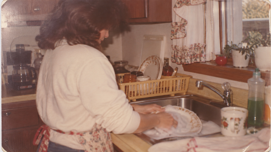 A person with brown hair washes dishes at a kitchen sink. Clean dishes are in a yellow rack, and a window with floral curtains is above the sink. Soap and a mug are on the counter.