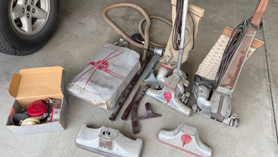 A collection of vintage Kirby vacuum cleaners, including two upright models, an attachment bag, various floor nozzles, a hose, and a box of small cleaning accessories.