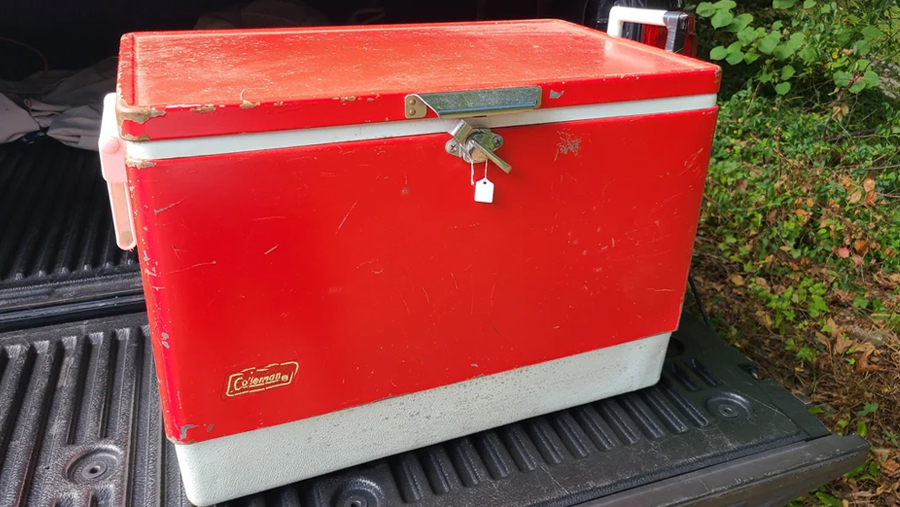 A vintage red and white Coleman cooler with a padlock on the lid sits on the bed of a black pickup truck, with green foliage in the background.