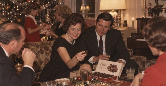 A group of adults in formal attire sit around a table sharing chocolates, with a decorated Christmas tree and children in the background. The setting is festive and warmly lit.