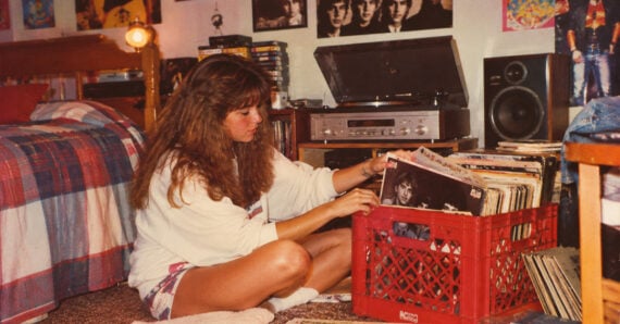 A woman sits on the floor by a crate of vinyl records in a cozy bedroom, flipping through albums. Nearby, a record player, speakers, and posters decorate the space, creating a vintage, retro vibe.