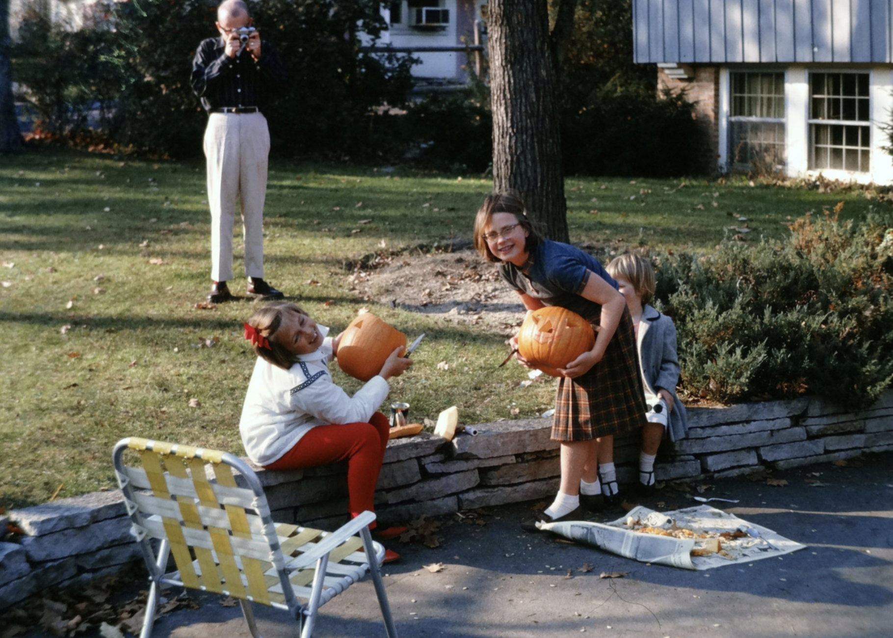 A man takes a photo of three children carving pumpkins on a stone wall in a yard.