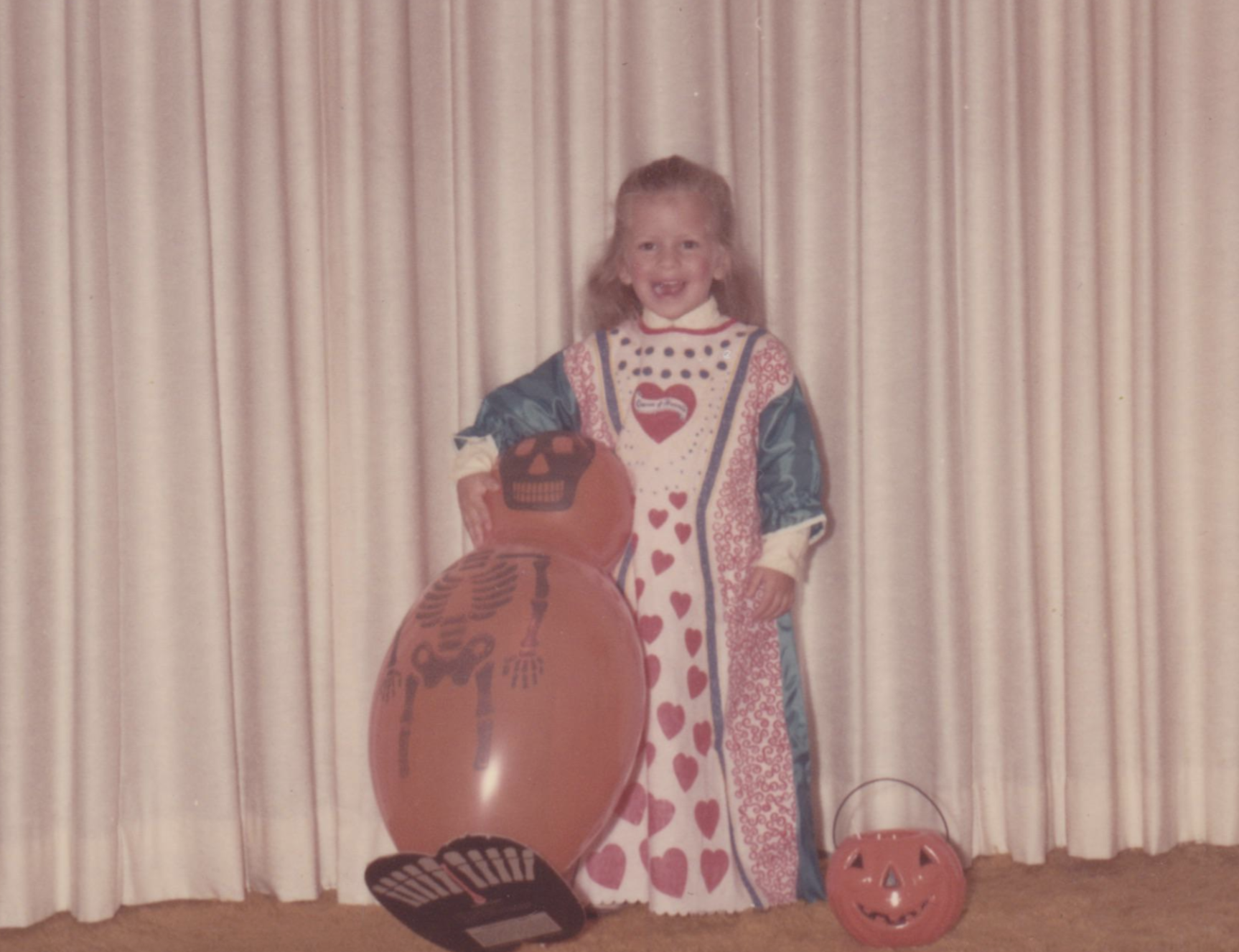 A young girl in a heart-patterned costume stands next to a large orange skeleton balloon and a jack-o’-lantern bucket.