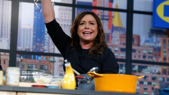 Rachael Ray smiling and raising her arm while cooking in a kitchen studio.