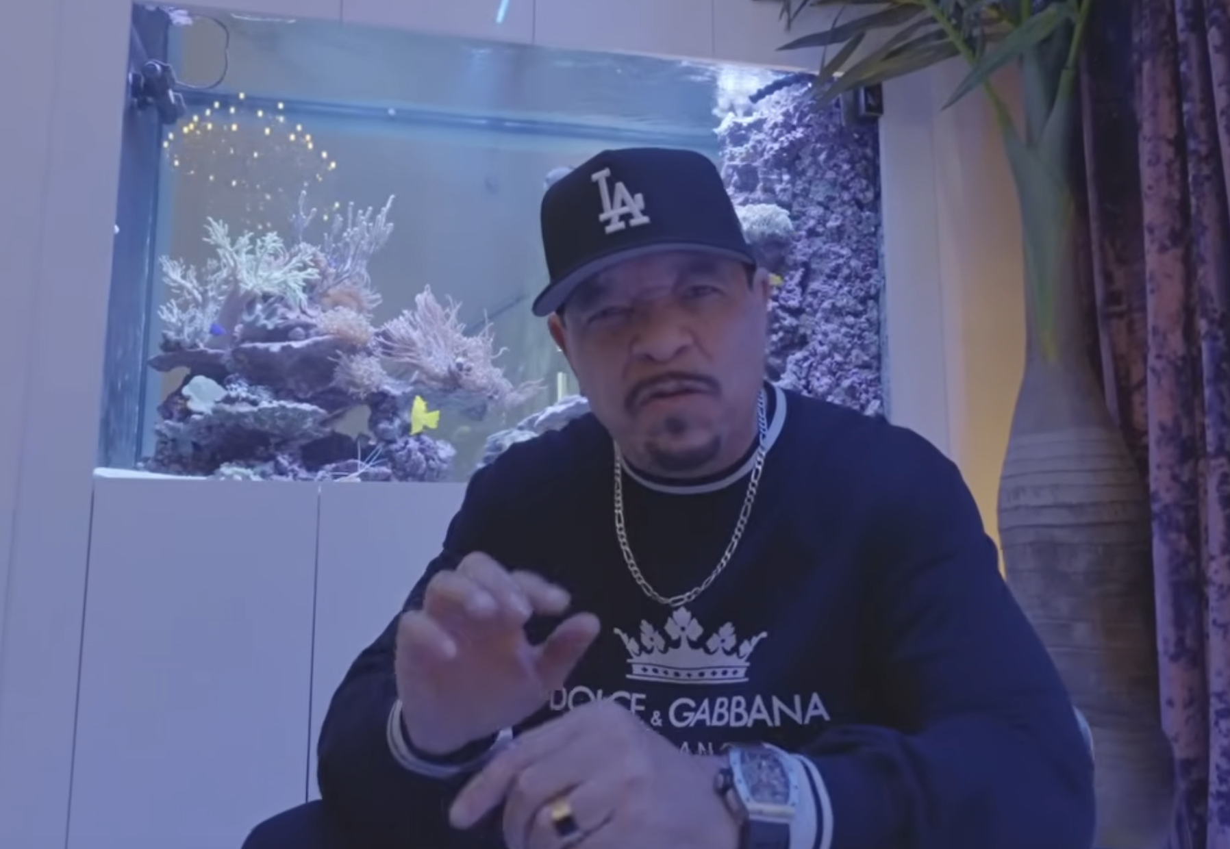 Ice-T wearing a black LA baseball cap and a black Dolce & Gabbana sweatshirt in front of a large aquarium.