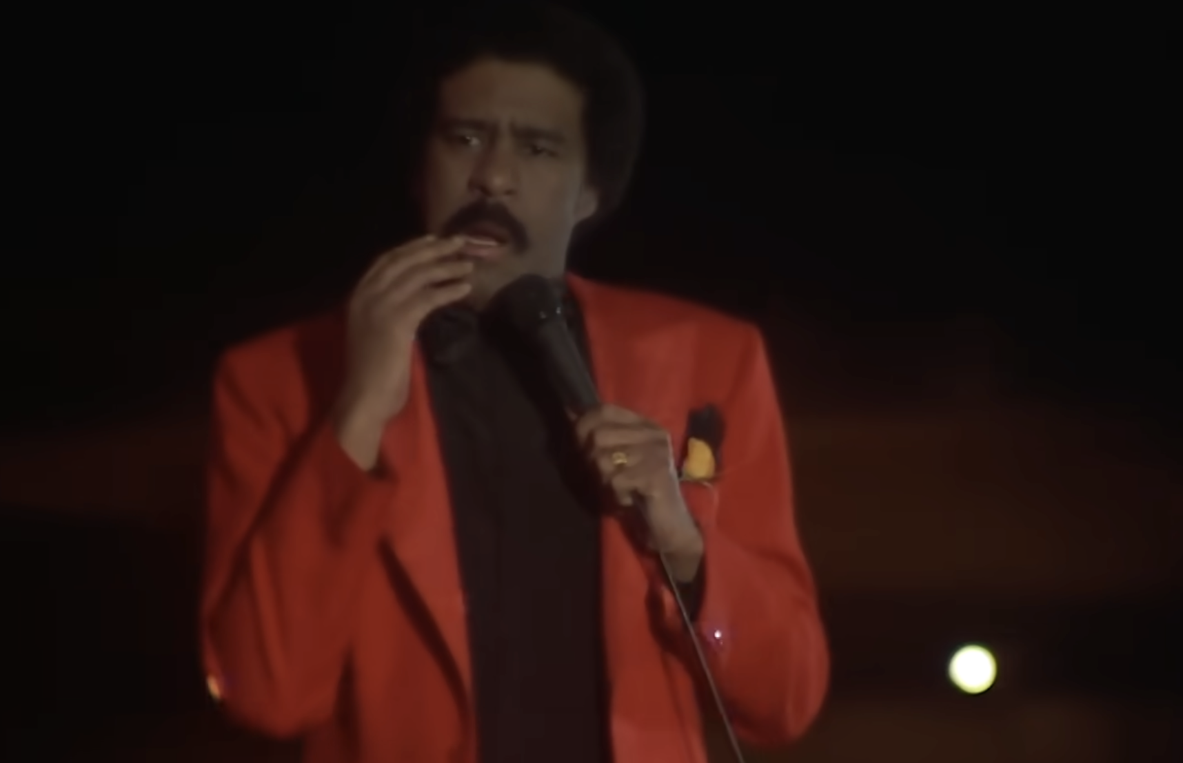 Richard Pryor performing on stage in a red blazer, holding a microphone.