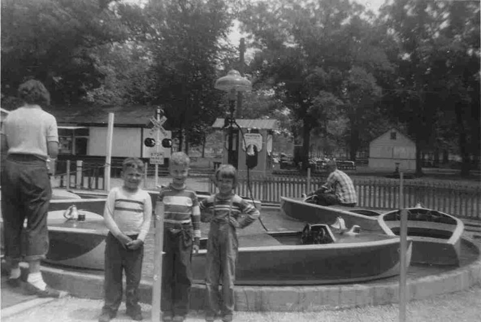 Three young boys stand in front of a small boat ride at a park. An adult stands to the left and another sits inside the ride area. Trees, a building, and a railroad crossing sign appear in the background. Black and white photo.