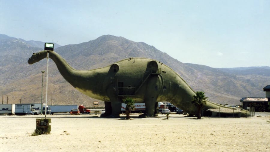 A giant dinosaur-shaped building standing in a desert landscape in front of a mountain range.