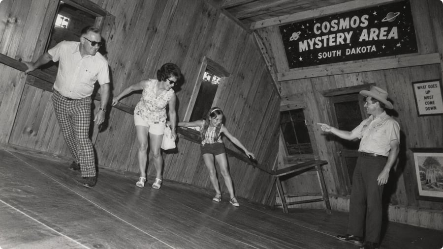 Three people leaning at an angle while walking through the slanted room of the Cosmos Mystery Area in South Dakota, as a man in a cowboy hat points toward them.