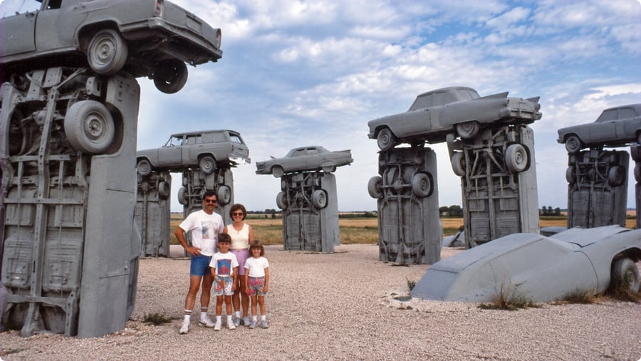 A family of four stands in front of Carhenge, a sculpture made of gray cars arranged to resemble Stonehenge.