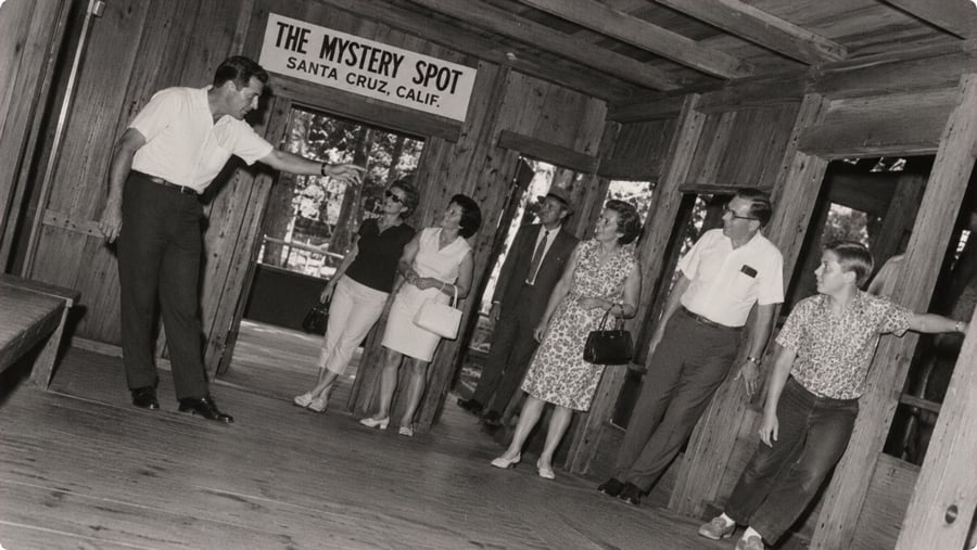 A guide points toward a sign for The Mystery Spot in Santa Cruz as a group of visitors stands on a tilted wooden floor.