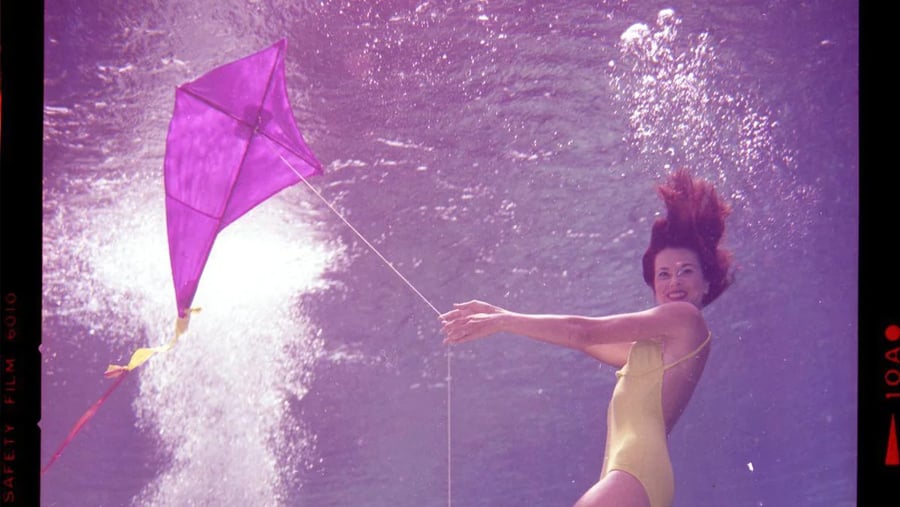 A woman in a yellow swimsuit swims underwater while holding the string of a purple kite.