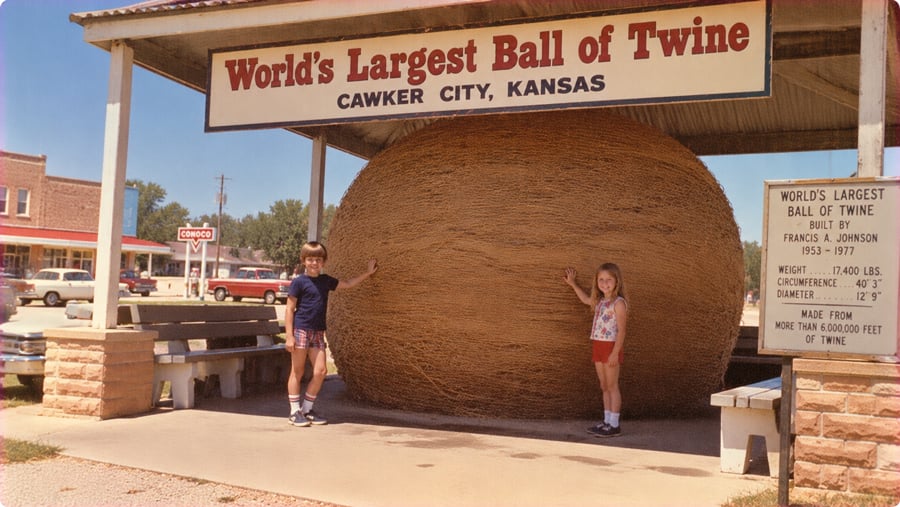 A young boy and girl stand on either side of the World’s Largest Ball of Twine in Cawker City, Kansas, with an informational sign nearby.