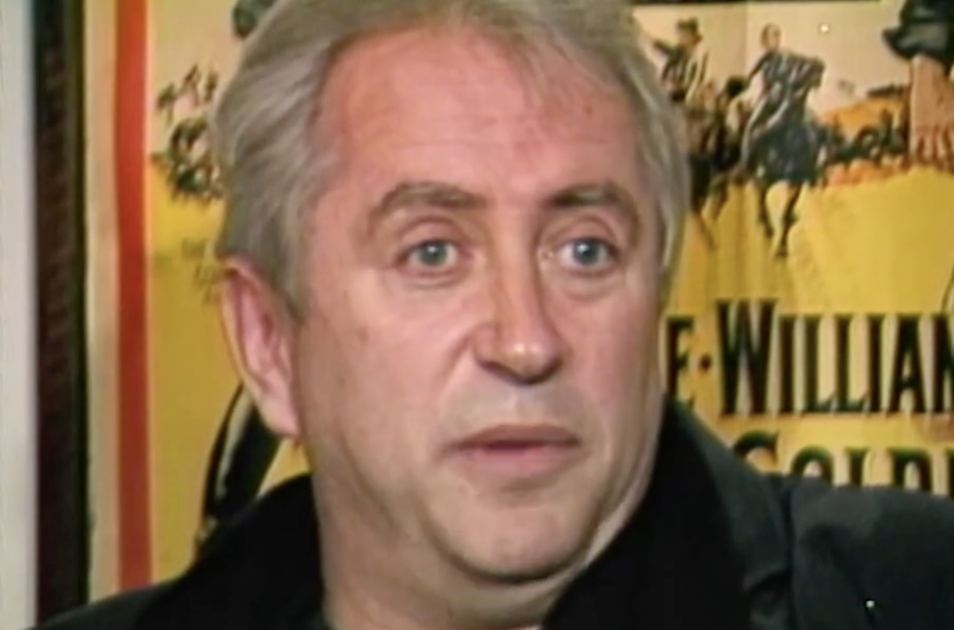A middle-aged man with short grey hair speaking in front of a vintage movie poster.
