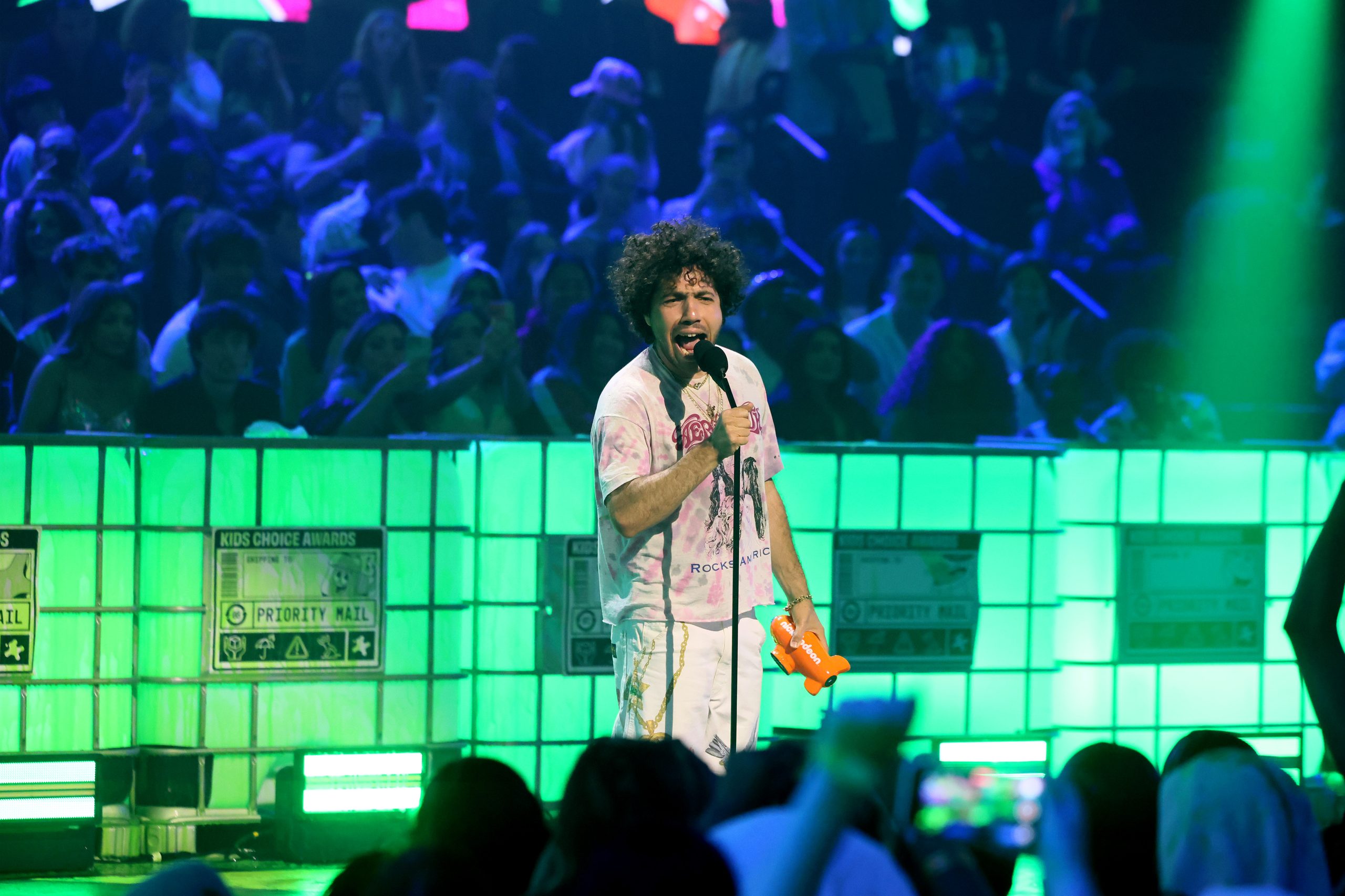 A performer with curly hair holding a microphone and an orange Nickelodeon blimp award on a stage lit with bright green lights, in front of a blurred audience.