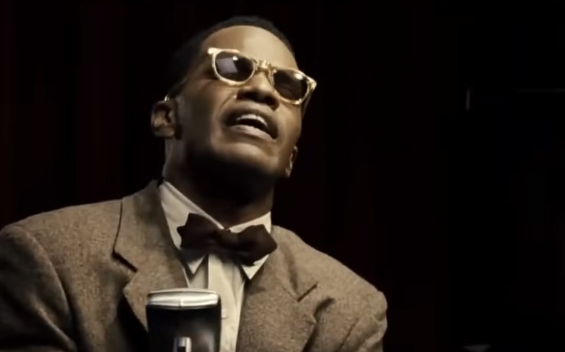 Jamie Foxx as Ray Charles singing into a microphone while wearing a suit, bow tie, and glasses.