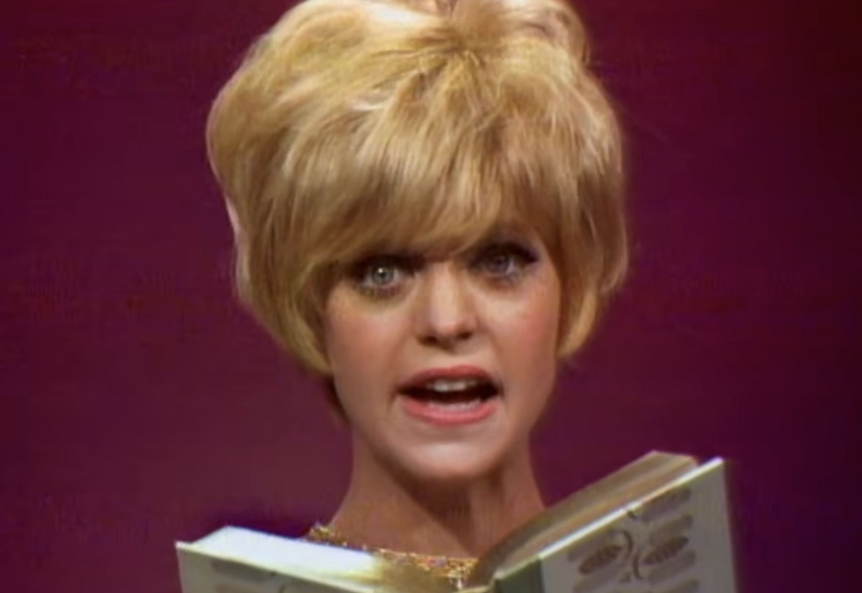 Goldie Hawn with short blonde hair holds an open book while looking at the camera.