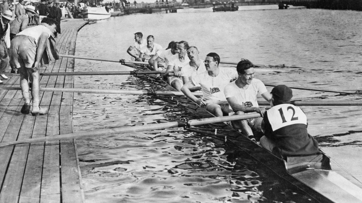 A rowing team of eight men in a boat, wearing numbered shirts, sits alongside a wooden dock with water and spectators in the background.
